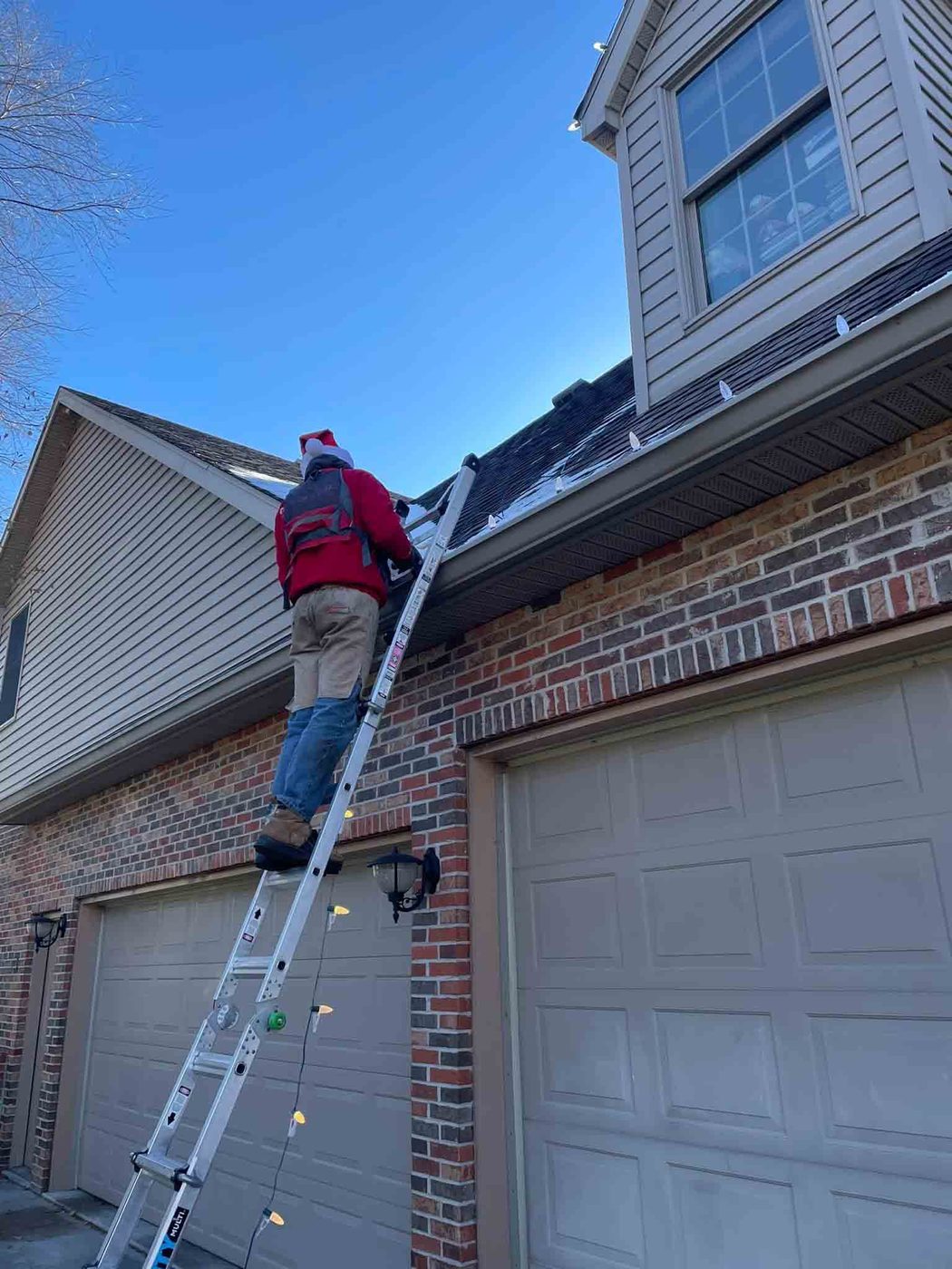 Indiana Christmas Light Installers crew member on a ladder fitting lights to a brick home under a clear blue sky