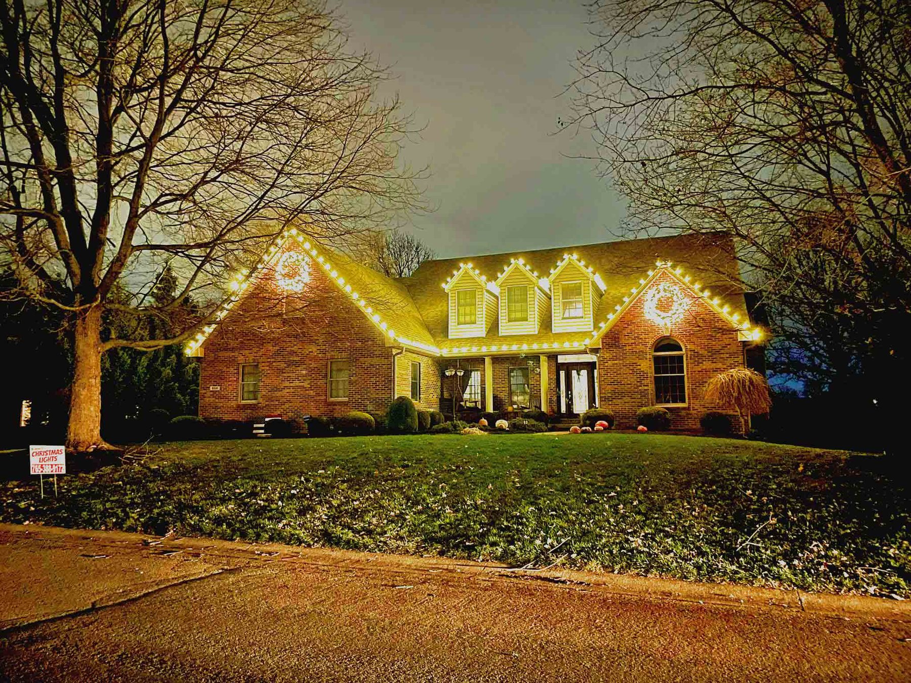 Brick Indiana home lit with warm white Christmas lights and large lighted wreaths on the gables
