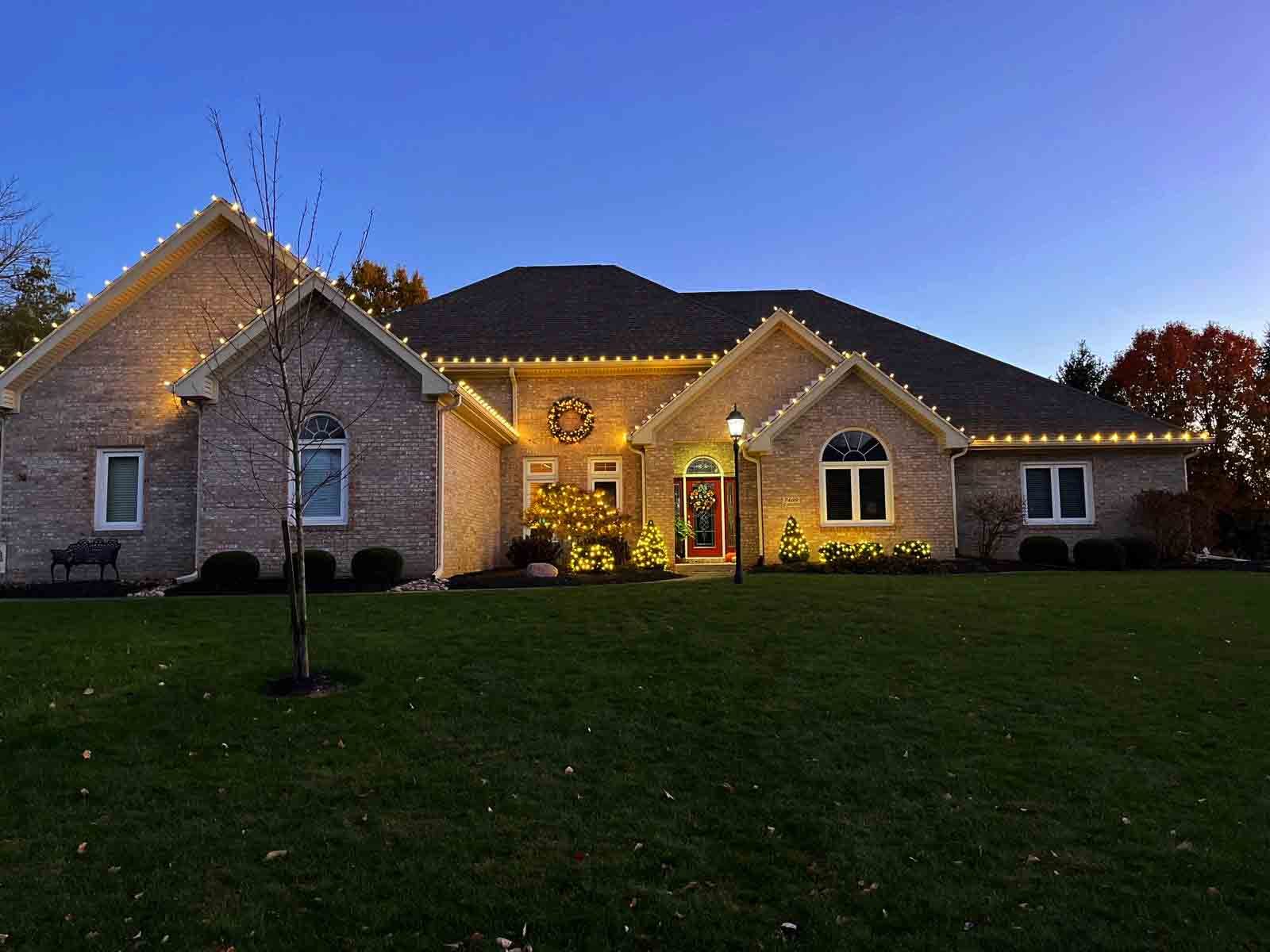 Craftsman style Indiana home outlined in warm white Christmas lights with a lighted gable wreath and lit shrubs along the front