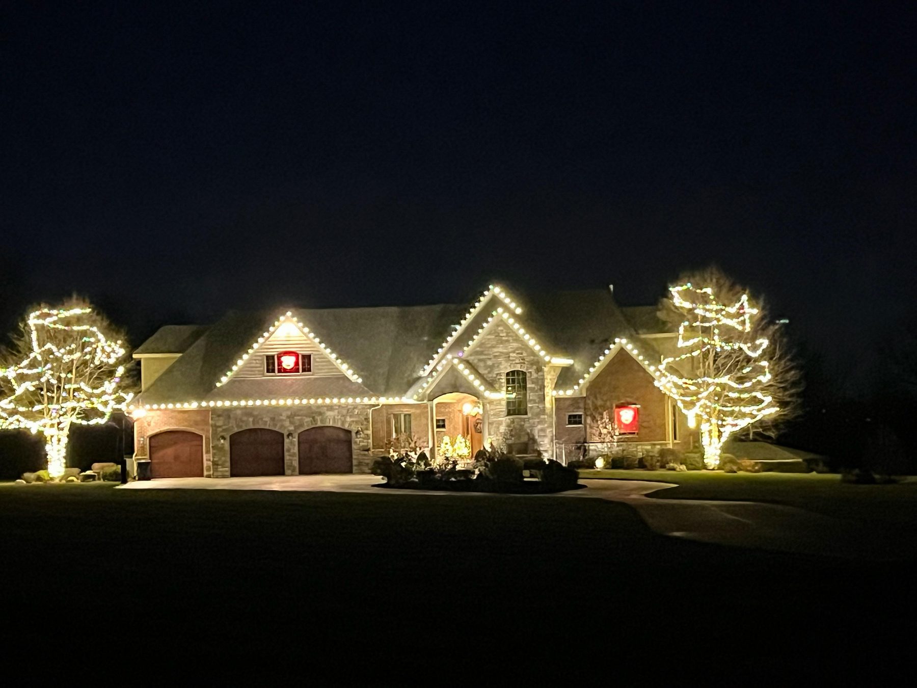 Large stone Indiana home with warm white Christmas lights on the rooflines, two mature trees wrapped trunk to branch and red lighted wreaths on the gables