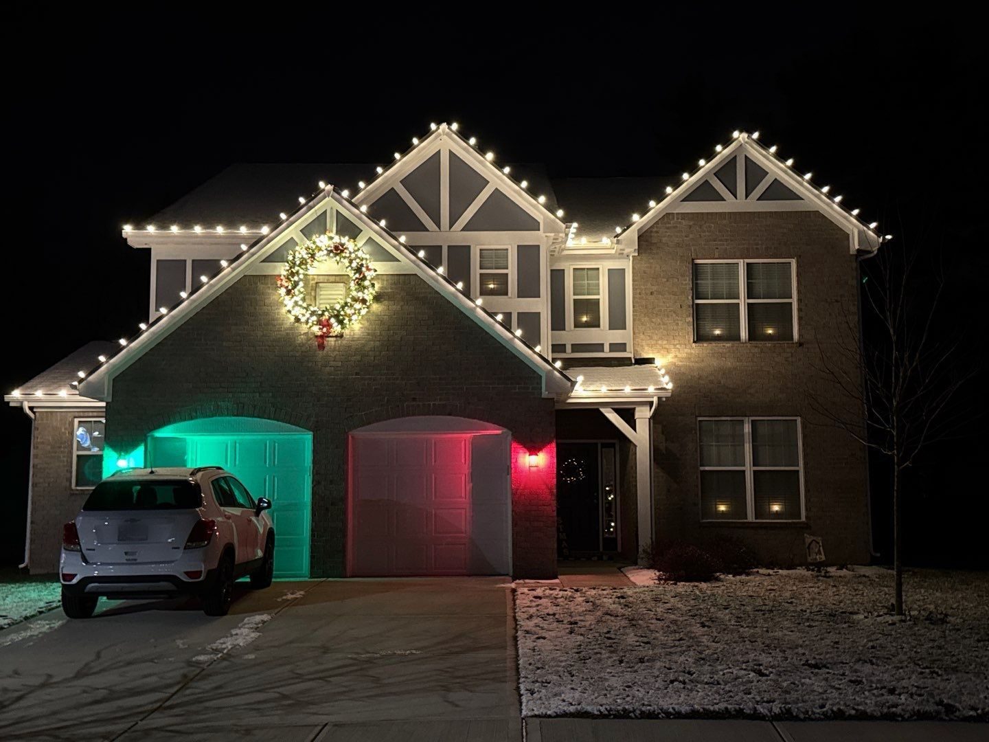 Two story Indiana home outlined in warm white Christmas lights with a large lighted wreath on the front gable and the garage front washed in red and green