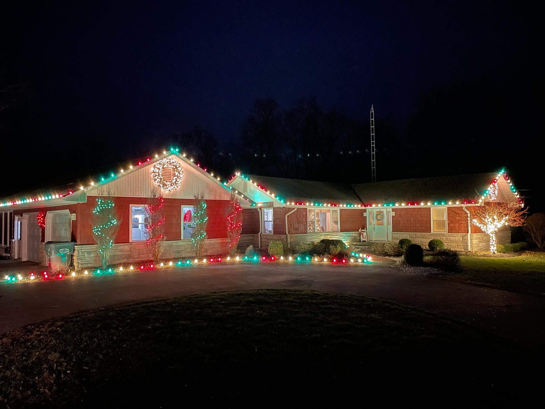 Long Indiana ranch home outlined in red and green Christmas lights with a lit gable wreath, lit shrubs and a wrapped tree