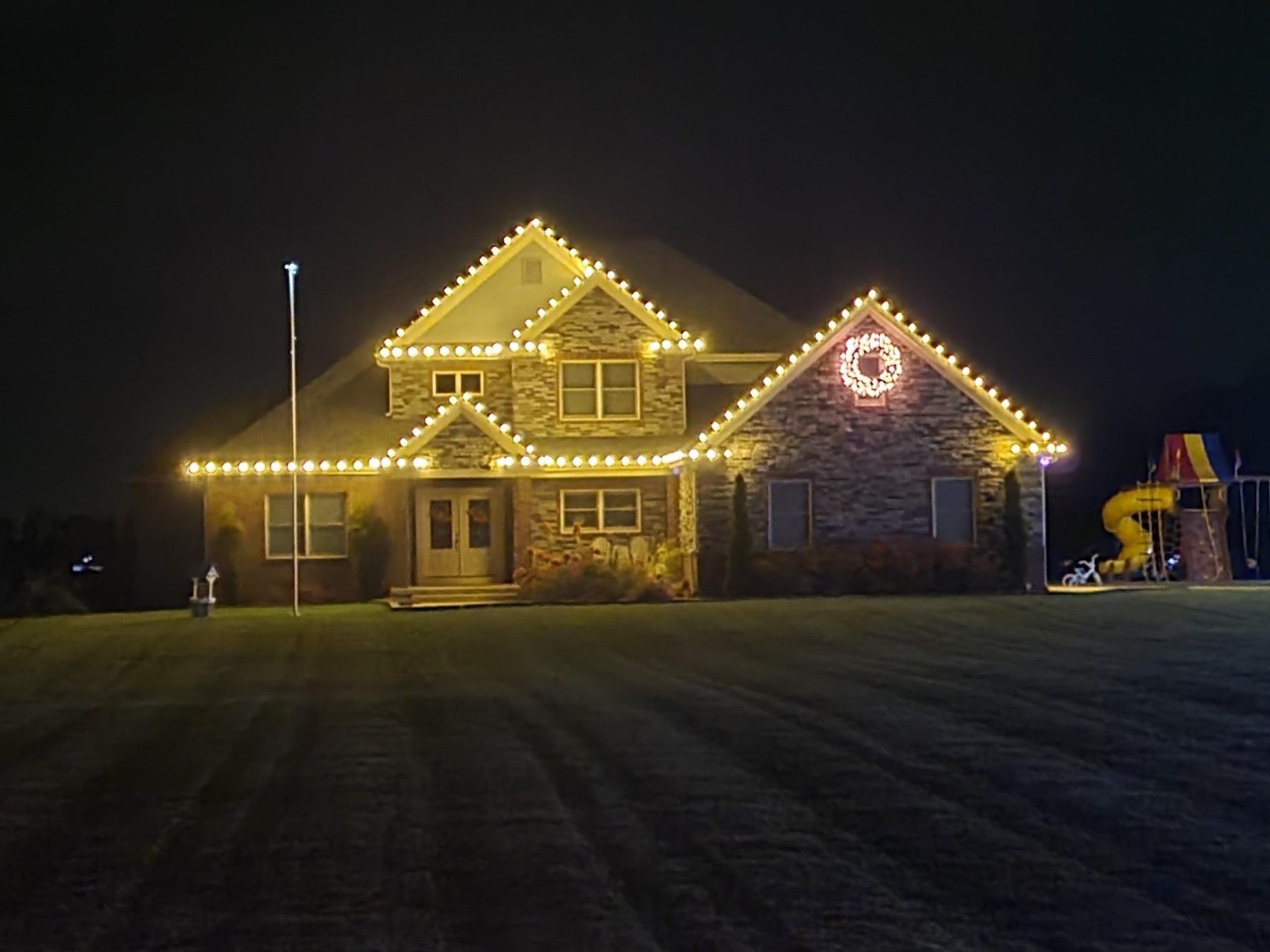 Stone Indiana home with warm white C9 Christmas lights along every gable and eave and a lighted wreath on the right gable