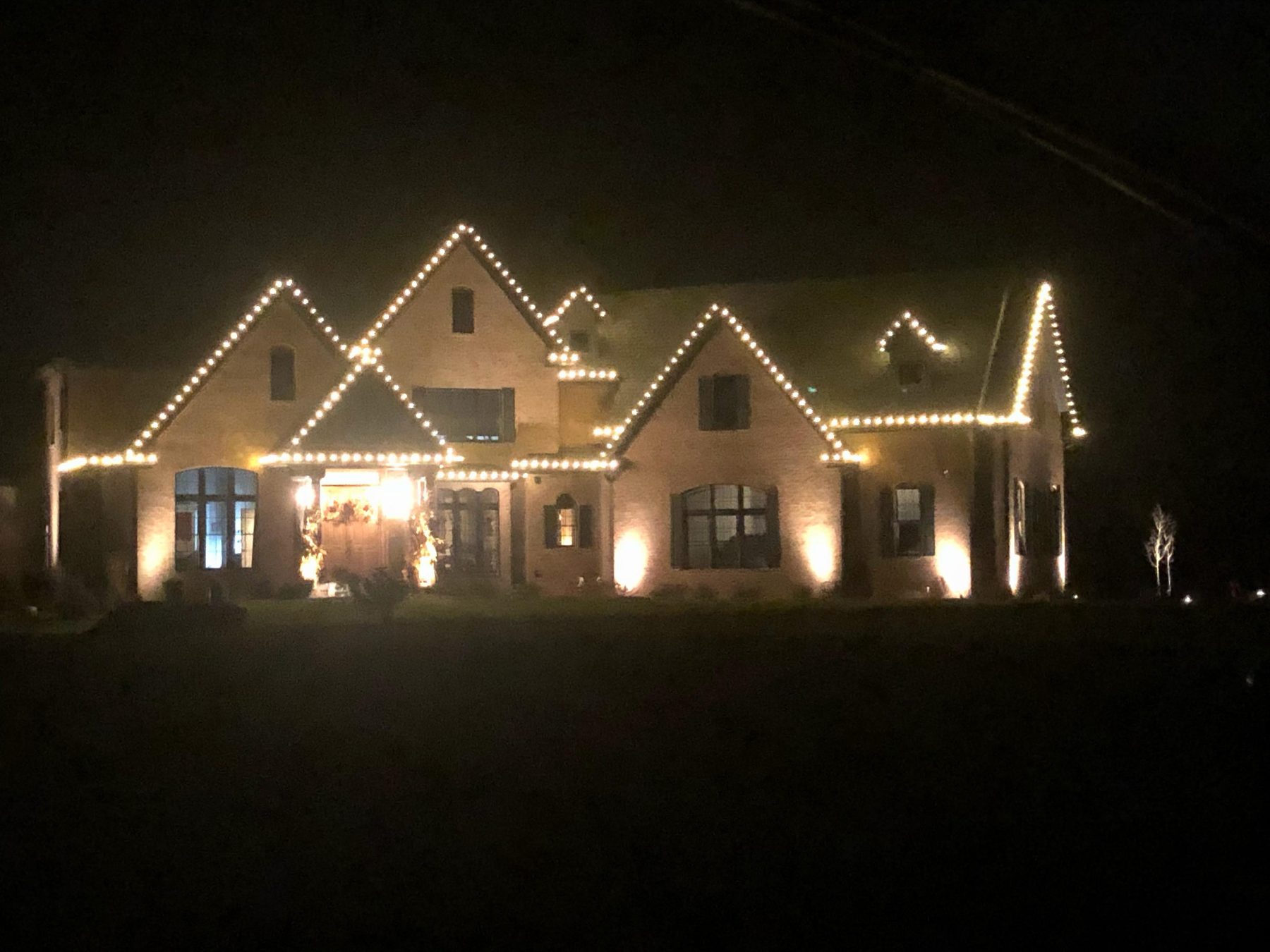 Indiana home with warm white Christmas lights along five roof peaks, uplighting across the brick and lit garland at the entrance
